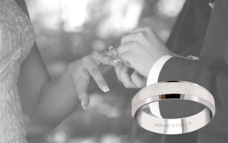 Pareja colocándose sus argollas de matrimonio. A un lado un ejemplo de argolla de matrimonio de oro blanco