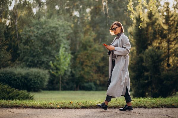 Mamá caminando al aire libre mientras revisa su celular, integrando movimiento ligero y autocuidado en su rutina diaria