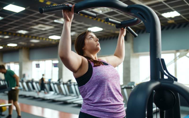 Mujer principiante utilizando máquina guiada de gimnasio para entrenar fuerza de brazos, siguiendo una rutina de ejercicio segura y controlada.