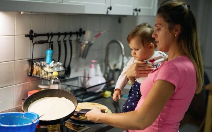 Madre multitaks cocina cargando un bebé en brazos, reflejo de la autoexigencia y carga mental invisible.