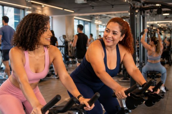 Dos mujeres entrenan en bicicleta fija en gimnasio moderno, imagen sobre mitos del ejercicio y evidencia científica.