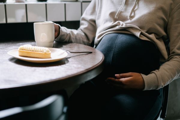 por que dan los antojos en el embarazo Mujer embarazada comiendo una tarta y una taza de café representando los antojos en el embarazo.